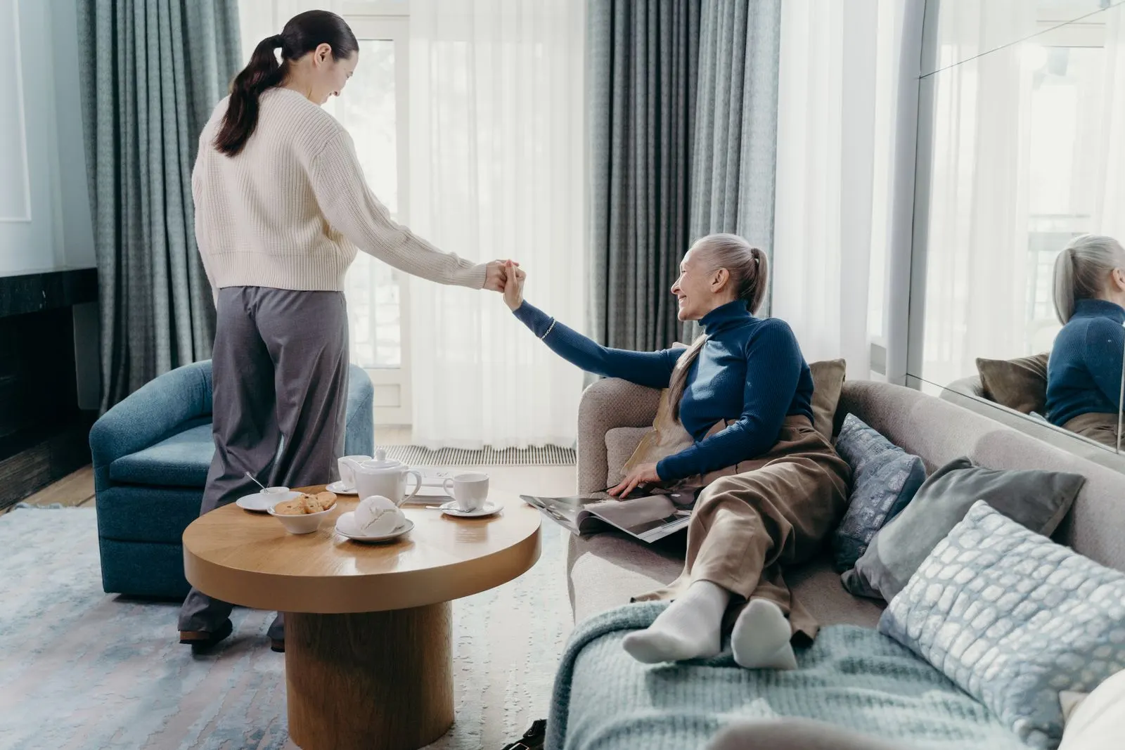An adult daughter helping her mother up from the sofa in a bright living room